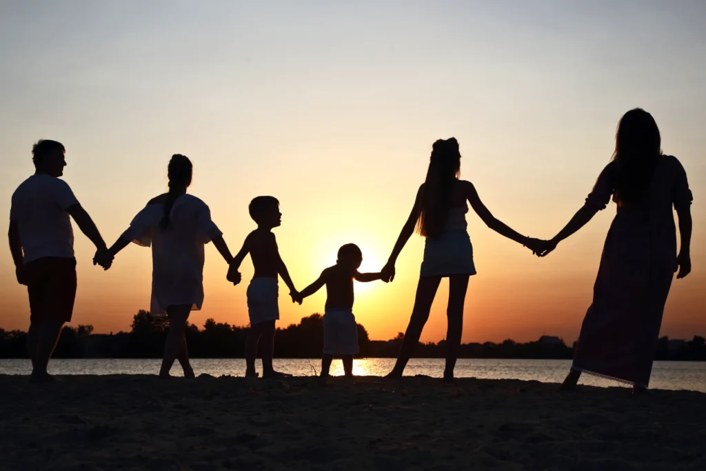 Silhouetten einer sechsköpfigen Familie, die bei Sonnenuntergang am Strand Händchen hält.